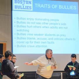 Teacher giving a Boston vs Bullies presentation and behind him is a presentation slide explaining the 'Traits of Bullies'